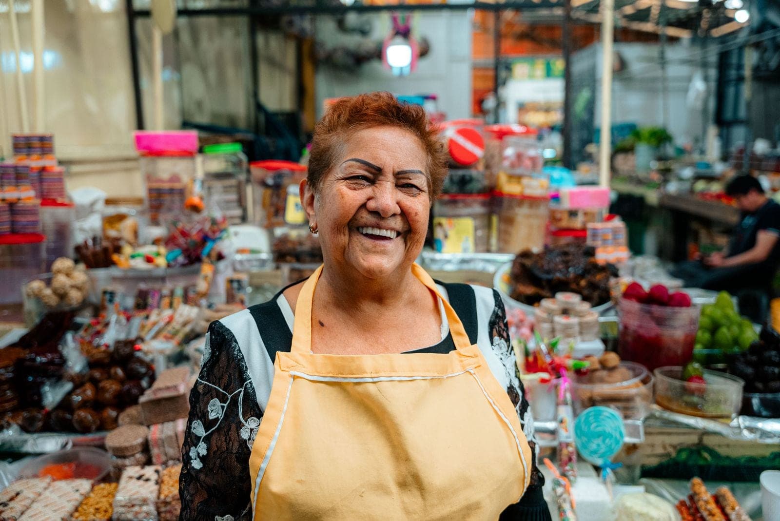 Comerciante sonríe frente al mostrador de su puesto de mercado, rodeada de dulces y productos de colores